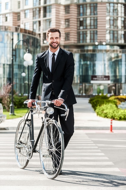 Der perfekte Weg zur Arbeit. In voller Länge lächelnder junger Geschäftsmann, der beim Überqueren der Straße sein Fahrrad rollt