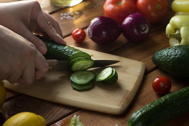 Der Prozess des Schneidens von Gemüse für vegetarischen Salat. Frauenhände schneiden Tomaten, Gurken, Zwiebeln, Paprika auf einem Holztisch.
