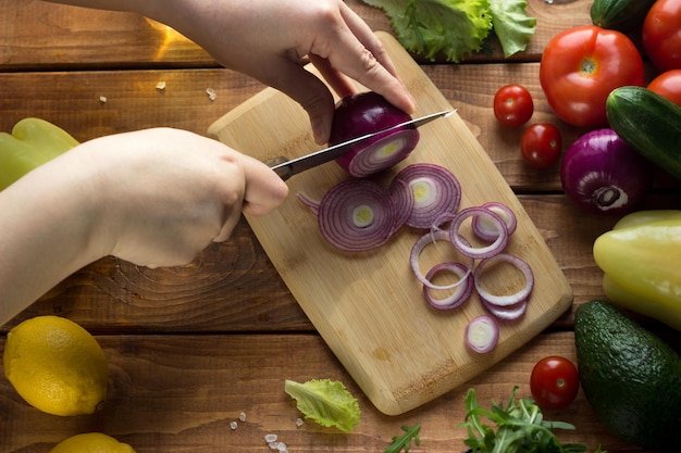 Der Prozess des Schneidens von Gemüse für vegetarischen Salat. Frauenhände schneiden Tomaten, Gurken, Zwiebeln, Paprika auf einem Holztisch.