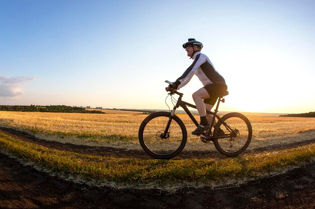 Der Radfahrer fährt mit dem Fahrrad auf der Straße in der Nähe des Feldes vor dem Hintergrund der untergehenden Sonne. Outdoor-Sportarten. Gesunder Lebensstil.