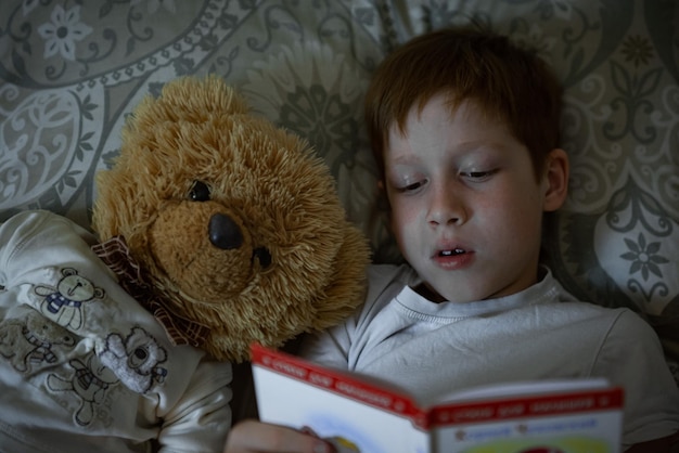 Der rothaarige Junge des Kindes liest ein Buch, bevor er ins Bett geht, liegt im Bett mit einem Teddybär-Nahaufnahmeporträt und lacht über den Lebensstil zu Hause