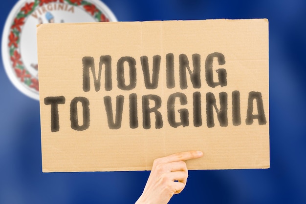 Der Satz "Moving to Virginia" auf einem Banner in Männerhand mit verschwommenem Meer im Hintergrund. Verlegung. Strand. Sonne. Entspannen. Warm. Wohnung. Umzug. Reisender