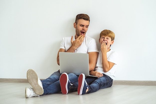 Der schläfrige Vater und ein Sohn mit einem Laptop sitzen auf dem Boden