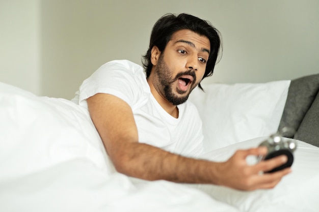 Der schockierte, gutaussehende, tausendjährige Mann aus dem Nahen Osten blickt mit offenem Mund auf den Wecker und liegt im Bett
