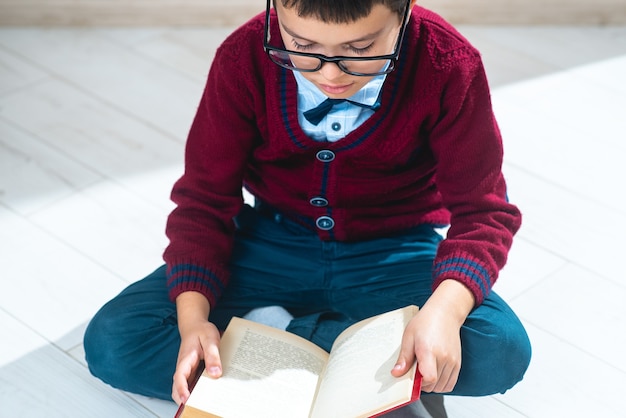 Der Schüler in Pullover und Brille sitzt im Lotussitz auf dem Boden und blättert in einem Buch