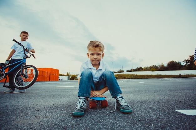 Der sechsjährige Junge sitzt auf dem Penny Board auf dem Parkplatz und schaut in die Kamera, sein älterer Bruder steht dahinter und hält sein Fahrrad. Porträt der aktiven Brüder