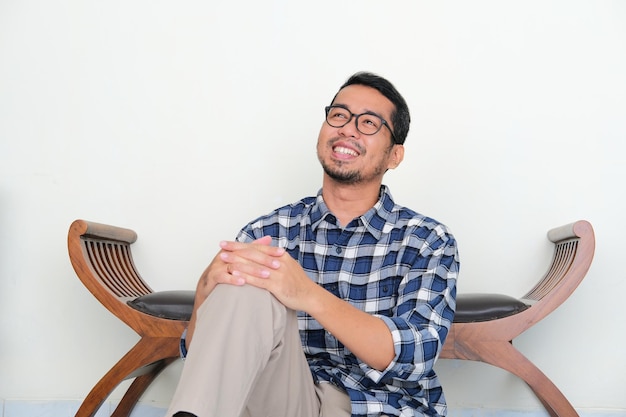 Der sitzende erwachsene asiatische Mann entspannt sich, während er sich etwas mit glücklichem Ausdruck vorstellt