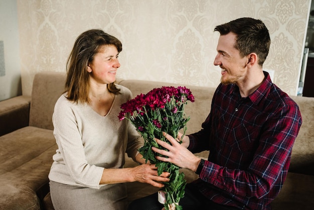 Der Sohn gibt ihrer Mutter Blumen und gratulierte zum Muttertag Portrait Vater und Großmutter lächeln und verbringen Zeit zusammen zu Hause Internationaler Frauentag Fröhliches Urlaubskonzept