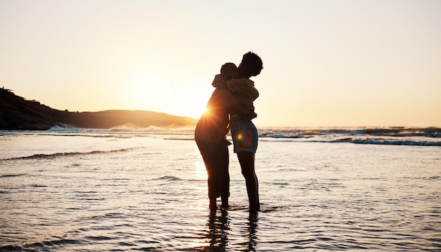 Der Sonnenuntergang am Strand und die Silhouette eines Paares, das sich umarmt, verbinden sich und genießen eine romantische Zeit zu zweit, verbinden sich und entspannen sich im Meerwasser. Dunkler Schatten, Sommerliebe oder Menschen im Freien, die sich auf tropischer Hochzeitsreise umarmen