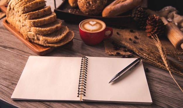 Der Stift wird auf ein weißes Notizbuch gelegt, das mit Kaffee und Brot auf einem Tisch ausgebreitet ist.
