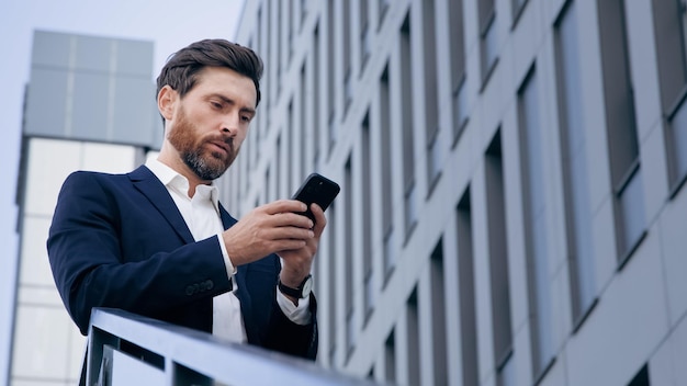 Der stilvolle, gutaussehende Geschäftsmann, der das Telefon hält und E-Mails liest, steht lächelnd auf der Straße und schreibt eine Nachricht