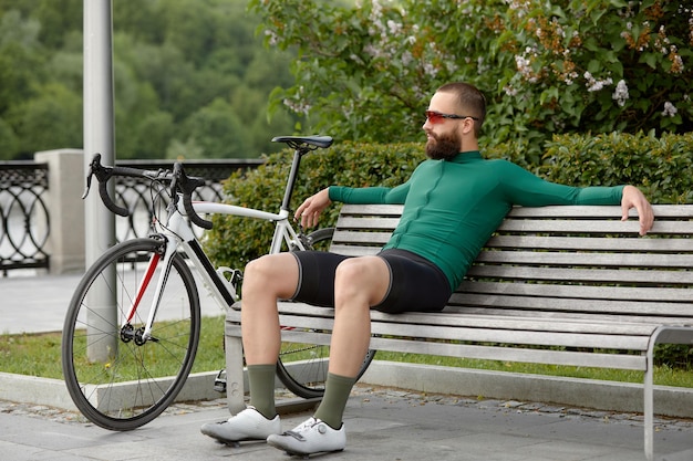 Der stilvolle, gutaussehende Radfahrer ruht auf einer Holzbank in der Nähe des Fahrrads im Sommerpark