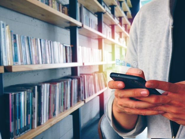 Der Student, der mit dem Handy nach neuen Medien sucht, während er in der Bibliothek einen Bericht erstellt