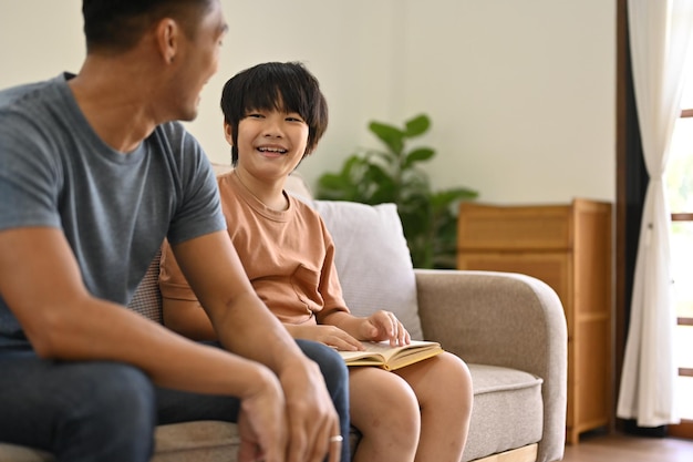 Der süße asiatische Junge sitzt mit seinem Vater im Wohnzimmer auf einem Sofa und verbringt Zeit mit der Familie