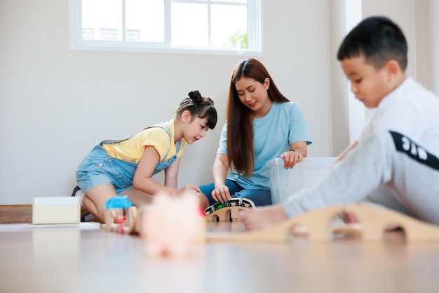 Der süße asiatische Junge spielt Holzblockbau-Eisenbahn und -Straße auf dem Boden, und seine Mutter und seine Schwester helfen ihm dabei, das zu Hause zu tun