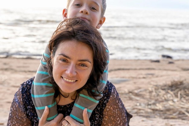 Der süße fröhliche Junge umarmt seine Mutter am Meer und schaut in die Kamera