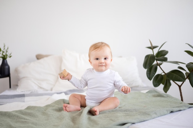 Der süße, gesunde Babyjunge in einem geräumigen, hellen Schlafzimmer sitzt auf einem Bett auf weißer Bettwäsche, das Kind lächelt Textilien für das Schlafzimmer und das Kinderzimmer