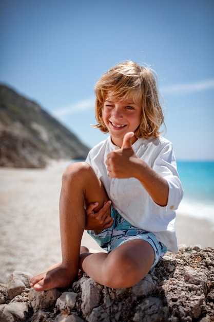 Der süße Junge sitzt auf dem Meeresfelsen am Strand, zeigt den Daumen nach oben und lächelt in die Kamera.