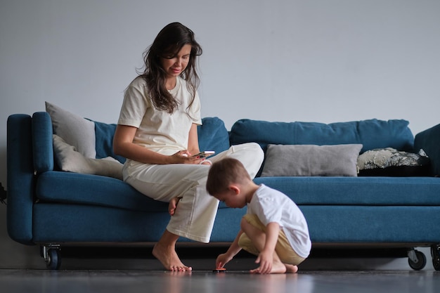 Der süße kleine Junge spielt auf dem Boden mit einem Auto, während die Mutter auf dem Sofa mit ihrem Smartphone surft