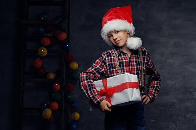 Der süße Teenager in Santas Hut, gekleidet in ein kariertes Hemd, hält eine große Geschenkbox.