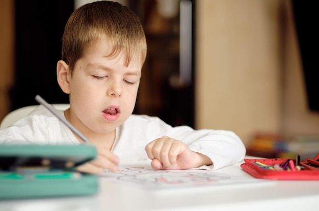 Der süße und lächelnde cucasian Junge zeichnet mit Buntstiften, während er am Tisch sitzt. Der Junge malt die Zeichnung mit einem roten Stift. Zeichenunterricht Kunsttherapie zum Stressabbau