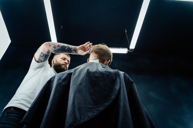 Der tätowierte Barber schneidet die Haare des Kunden mit einer Schere aus der Nähe Attraktiver Mann bekommt im Friseursalon einen modernen Haarschnitt Ansicht von unten