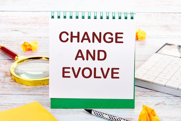 Der Text „CHANGE AND EVOLVE“ auf dem weißen Blatt des Notizbuchs befindet sich neben Aufklebern mit Lupe, Taschenrechner und Bleistift