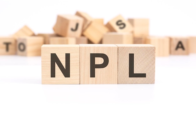 Der Text NPL Non Performing Loans steht auf drei Holzwürfeln, die auf einem weißen Tisch stehen, im Hintergrund ein Berg aus Holzwürfeln mit Buchstaben