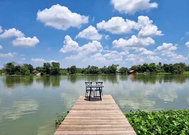 Der Tisch und die Stühle auf einer Holzbrücke landen am Flussufer und Wolken im Hintergrund des blauen Himmels