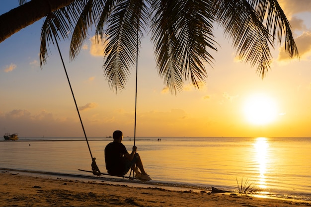 Der Typ genießt den Sonnenuntergang auf einer Schaukel am tropischen Strand.