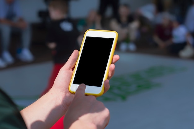 Der Typ hält vor dem Hintergrund der Kindertanzmeisterschaft ein Smartphone-Modell in der Hand.