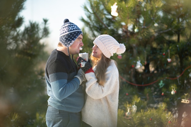 Der Typ mit dem Mädchen geht spazieren und küsst sich im Winterwald mit einer Tasse heißem Getränk. Ein gemütlicher Winterspaziergang durch den Wald mit einem heißen Getränk. Liebespaar, Winterurlaub