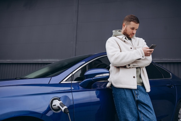 Der Typ setzte sich auf die Motorhaube seines Autos Sein Auto lädt an der Ladestation Ein Mann schaut auf den Bildschirm des Smartphones und lächelt