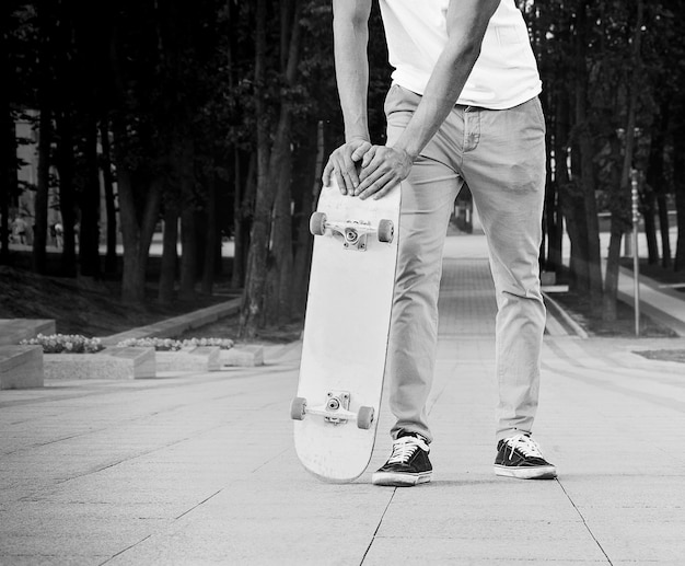 Der Typ steht im Park in der Sonne und lehnt sich an sein Skateboard