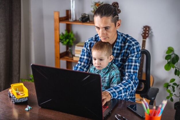 Der Vater eines jungen modernen Mannes arbeitet an einem Laptop, und sein kleiner Sohn sitzt auf seinem Schoß