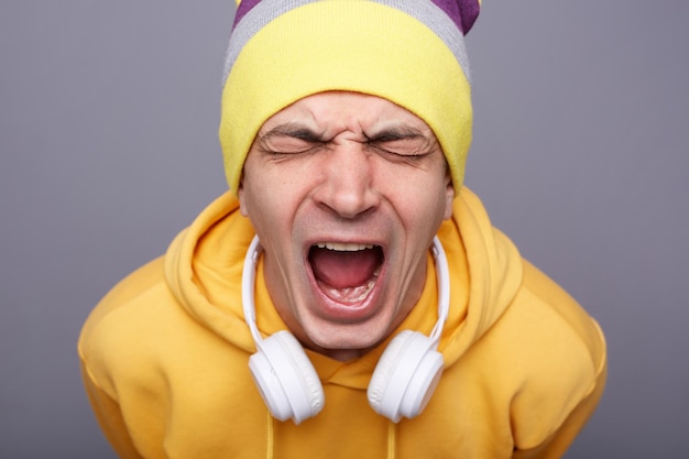 Der von Verzweiflung gestresste Hipster-Mann mit gelbem Hoodie und Mütze hält die Augen geschlossen und schreit laut, weil er depressiv ist. Er hat ernsthafte Probleme, an einer grauen Wand zu stehen und negative Emotionen auszudrücken