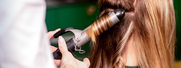 Der weibliche Friseur lockt Haare für eine braunhaarige junge kaukasische Frau in einem Schönheitssalon