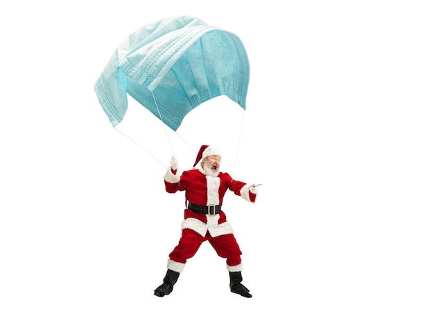 Der Weihnachtsmann fliegt auf einer riesigen Gesichtsmaske wie auf einem Ballon, der auf weißem Hintergrund isoliert ist. Kaukasisches männliches Modell in traditioneller Tracht. Neujahr, Geschenke, Feiertage, Winter, COVID, Pandemiekonzept. Exemplar.