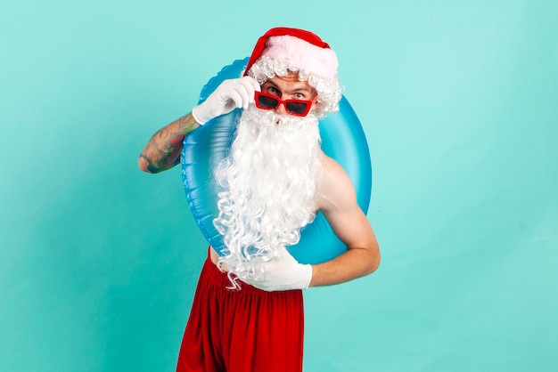 Der Weihnachtsmann im Anzug mit aufblasbarem Schwimmring und Sonnenbrille ruht am Strand auf blauem Hintergrund