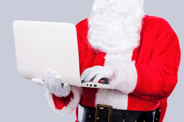 Der Weihnachtsmann im digitalen Zeitalter. Der Weihnachtsmann arbeitet am Laptop, während er vor grauem Hintergrund steht