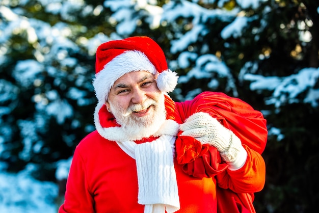 Der Weihnachtsmann kommt mit einer Tüte Geschenke auf der Schneelandschaft in den Winterwald. Der Weihnachtsmann trägt den Kindern an Heiligabend Geschenke in einer roten Tasche.