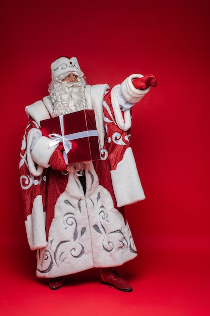 Der Weihnachtsmann mit dem langen weißen Bart zeigt etwas mit seiner Hand und hält ein Geschenk, Bild isoliert auf roter Wand