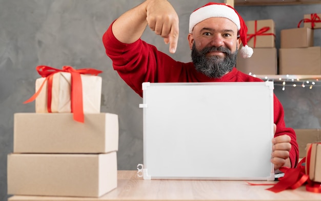 Der Weihnachtsmann mit roter Mütze arbeitet im Heimbüro und zeigt mit dem Finger auf eine Tafel mit einem Platz für den Text. Kleinunternehmer-Verkäufer für Weihnachtsprodukt-Werbekonzept