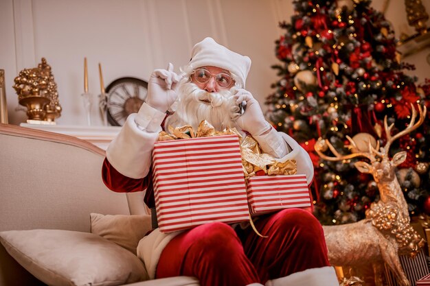 Der Weihnachtsmann sitzt auf der Couch und telefoniert mit dem Handy in der Nähe des Kamins und des Weihnachtsbaums mit Geschenken. Neues Jahr und frohe Weihnachten, frohes Feiertagskonzept