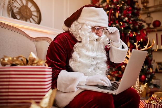 Der Weihnachtsmann sitzt bei ihm zu Hause und liest E-Mails auf einem Laptop mit Weihnachtsanfragen oder Wunschliste in der Nähe des Kamins und des Baumes mit Geschenken.
