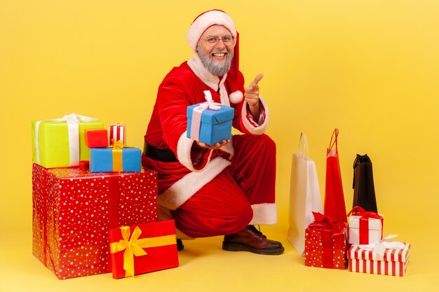 Der Weihnachtsmann sitzt in der Nähe von verpackten Geschenkboxen, zeigt auf die Kamera und gibt Geschenk und lächelt.