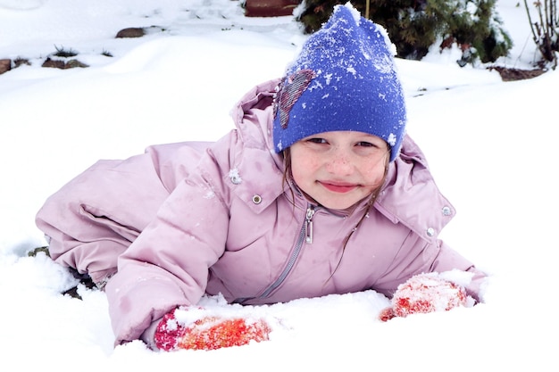 Der Winter ist eine Zeit des Spiels und Vergnügens. Ein achtjähriges lächelndes Mädchen in einer Pastelljacke und einem blauen Hut macht Schneebälle in einem verschneiten Hof