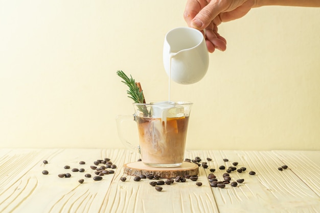 Derramando leite em um copo de café preto com cubo de gelo, canela e alecrim na superfície de madeira