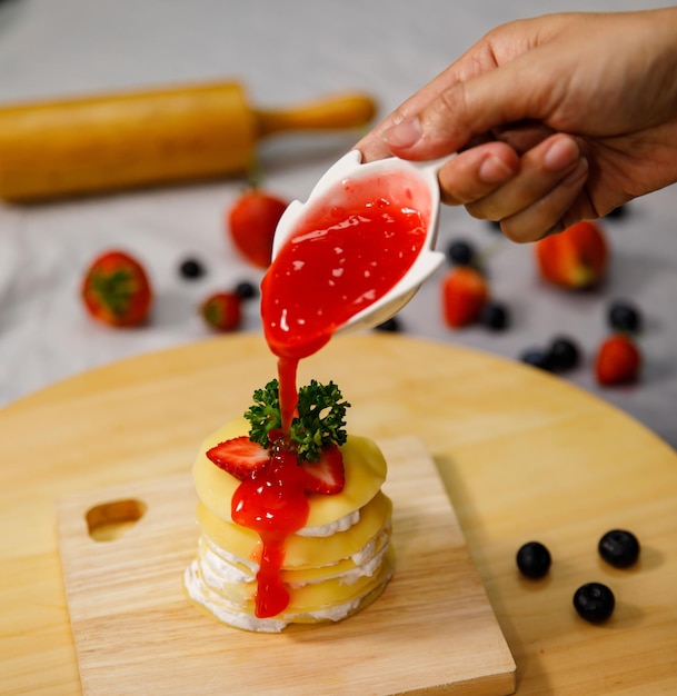 Derramando molho de morango no delicioso bolo de crepe, decorado com morangos frescos em uma placa de madeira, para aniversário, dia dos namorados e celebração