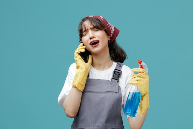 Desagradado jovem limpador feminino vestindo bandana uniforme e luvas de borracha segurando limpador falando no telefone com os olhos fechados, isolados no fundo azul